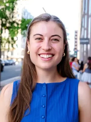 A woman in a blue shirt with long brown hair, smiling on a city street.
