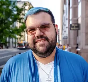 Portrait of a young man with glasses, a beard, and dark hair with blue highlights, smiling in front of a city street.
