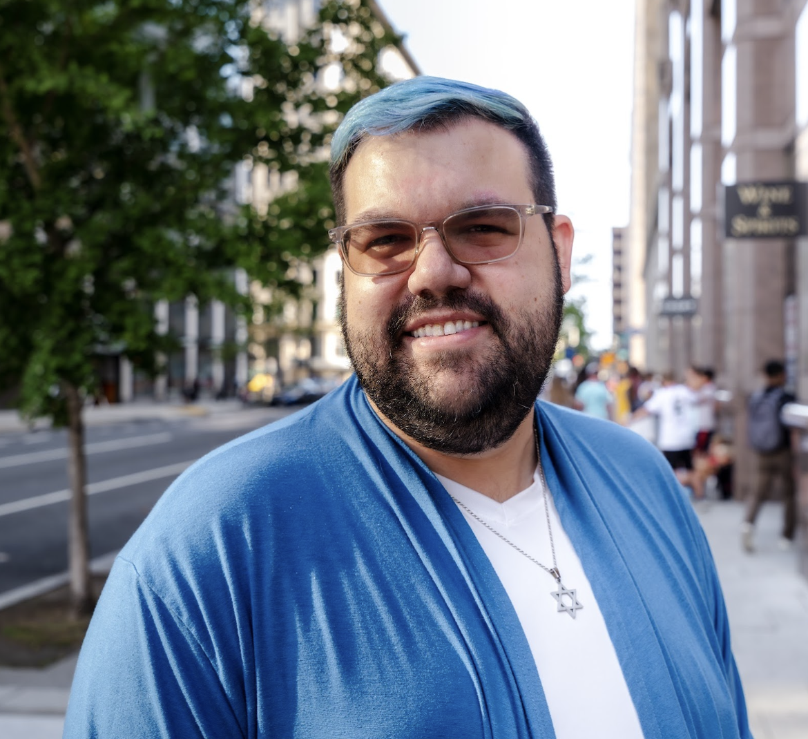 A smiling man with glasses, a beard, and blue hair, wearing a blue jacket and a Star of David necklace, standing on a city sidewalk with trees and buildings in the background.
