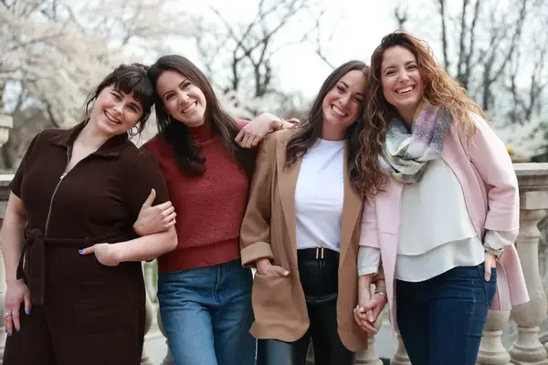 Four women standing outdoors, smiling, with trees in the background.