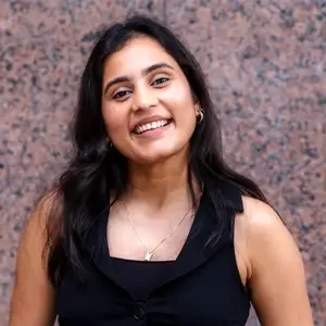 A smiling woman with long dark hair wearing a black sleeveless top, gold hoop earrings, a gold necklace, and standing in front of a speckled brown wall.