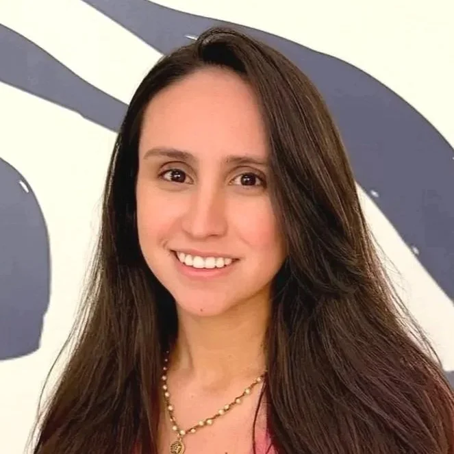 A woman with long brown hair, wearing a pink top and a beaded necklace, smiling at the camera.