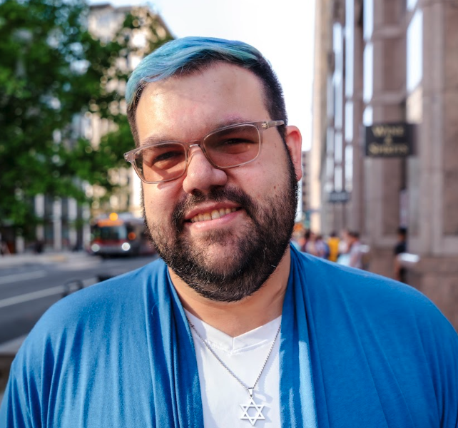 Portrait of a young man with glasses, a beard, and dark hair with blue highlights, smiling in front of a city street.