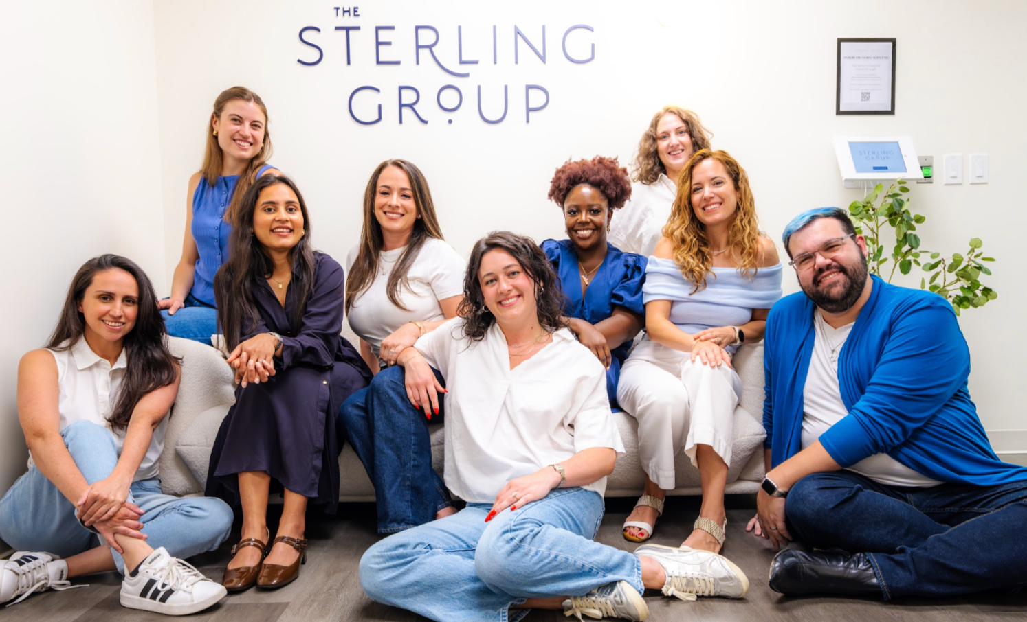 A diverse group of people, all in various shades of white and blue, sitting together on a couch and the floor underneath a sign that says "The Sterling Group."