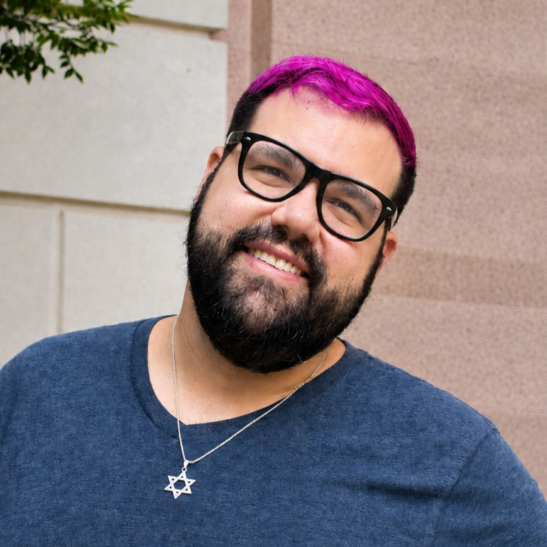 A man with a beard, wearing clear glasses and a blue shirt, smiling against a city street background. He has colorful blue hair.