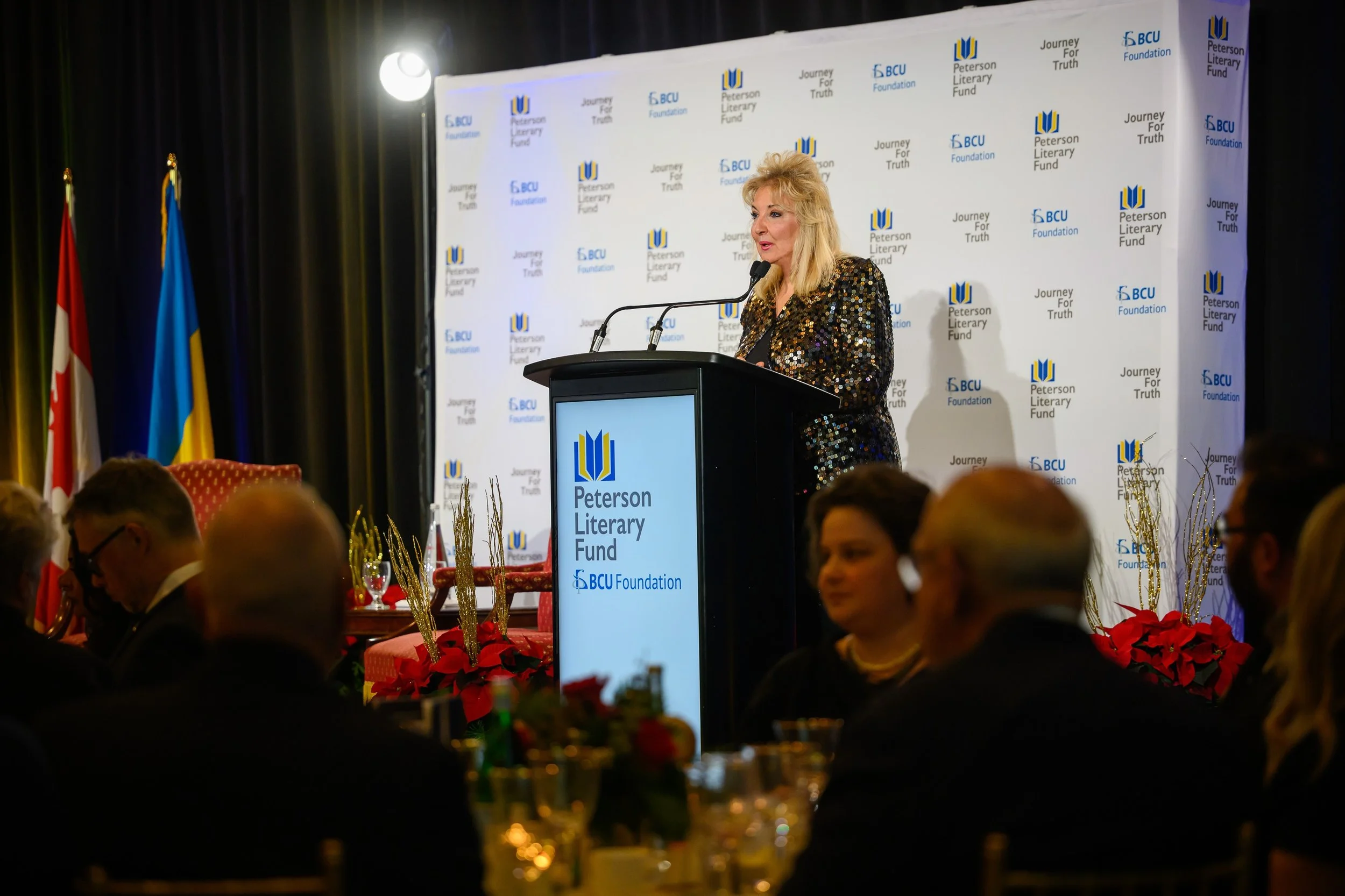 A woman with blonde hair speaking at a podium during an event, with a backdrop displaying the Peterson Literary Fund and BCU Foundation logos, and flags on the side.