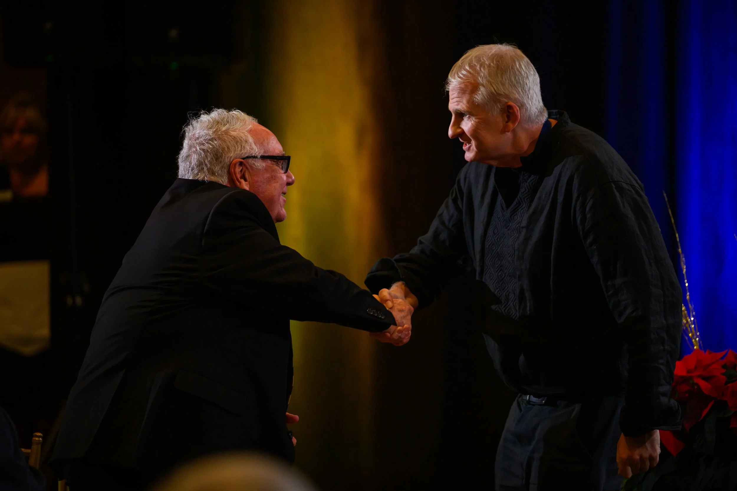 Two men, one older with gray hair and glasses and the other younger with blond hair, shaking hands during a formal event.