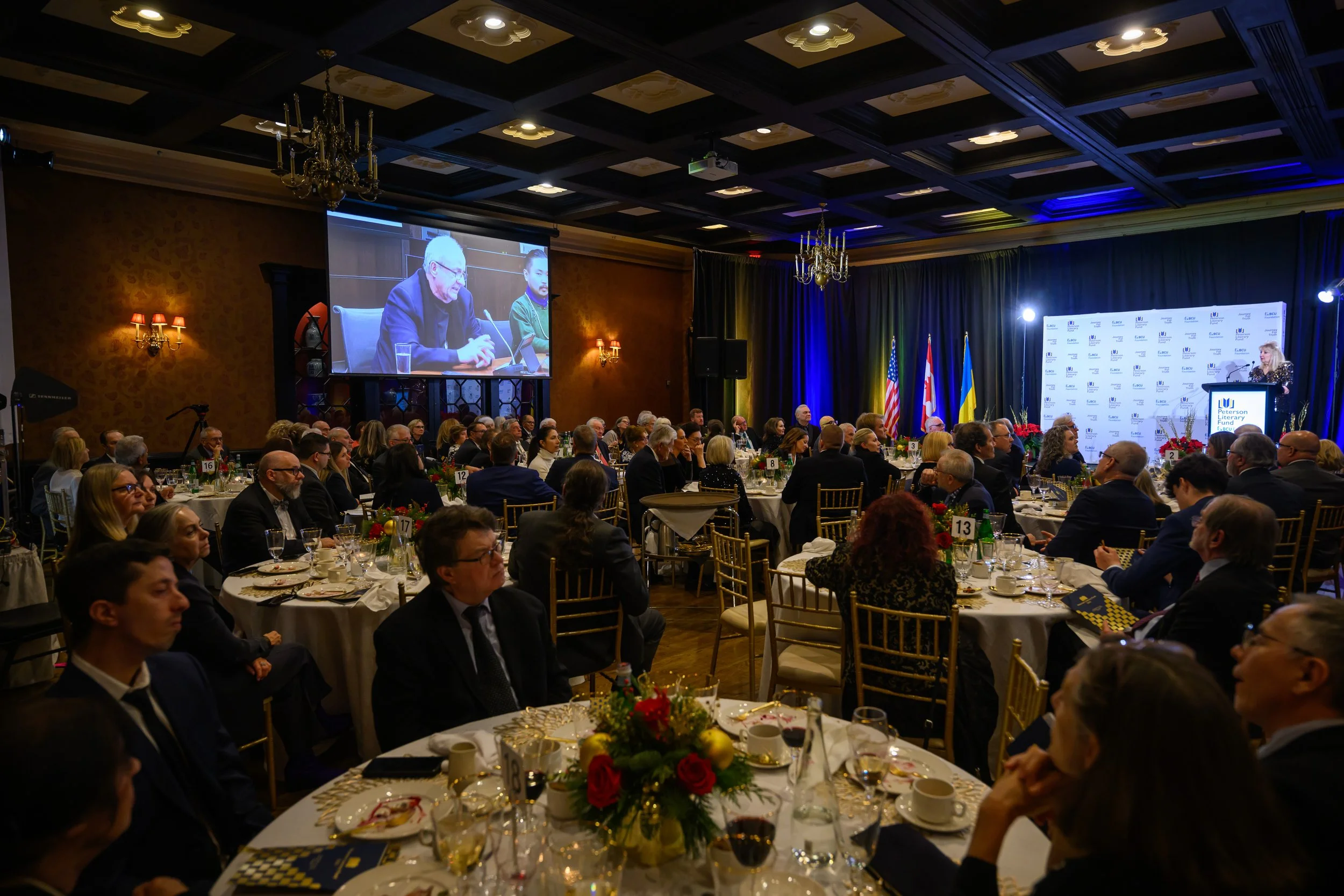 A large formal banquet hall filled with round tables dressed with white tablecloths, decorated with floral centerpieces and holiday-themed accents. Many people are seated, facing a stage with multiple large screens displaying a speaker. The stage background features flags of the United States, the United Kingdom, and Ukraine, and the venue has ornate chandeliers and wall sconces providing warm lighting.