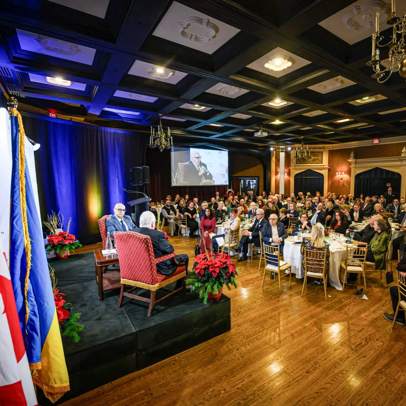 A conference or formal event with a speaker on a stage, an audience seated at round tables, a large screen displaying a man speaking into a microphone, and festive decorations including poinsettias and poinsettia-themed centerpieces. The room has dark wood walls, ornate chandeliers, and a dark ceiling with decorative panels.