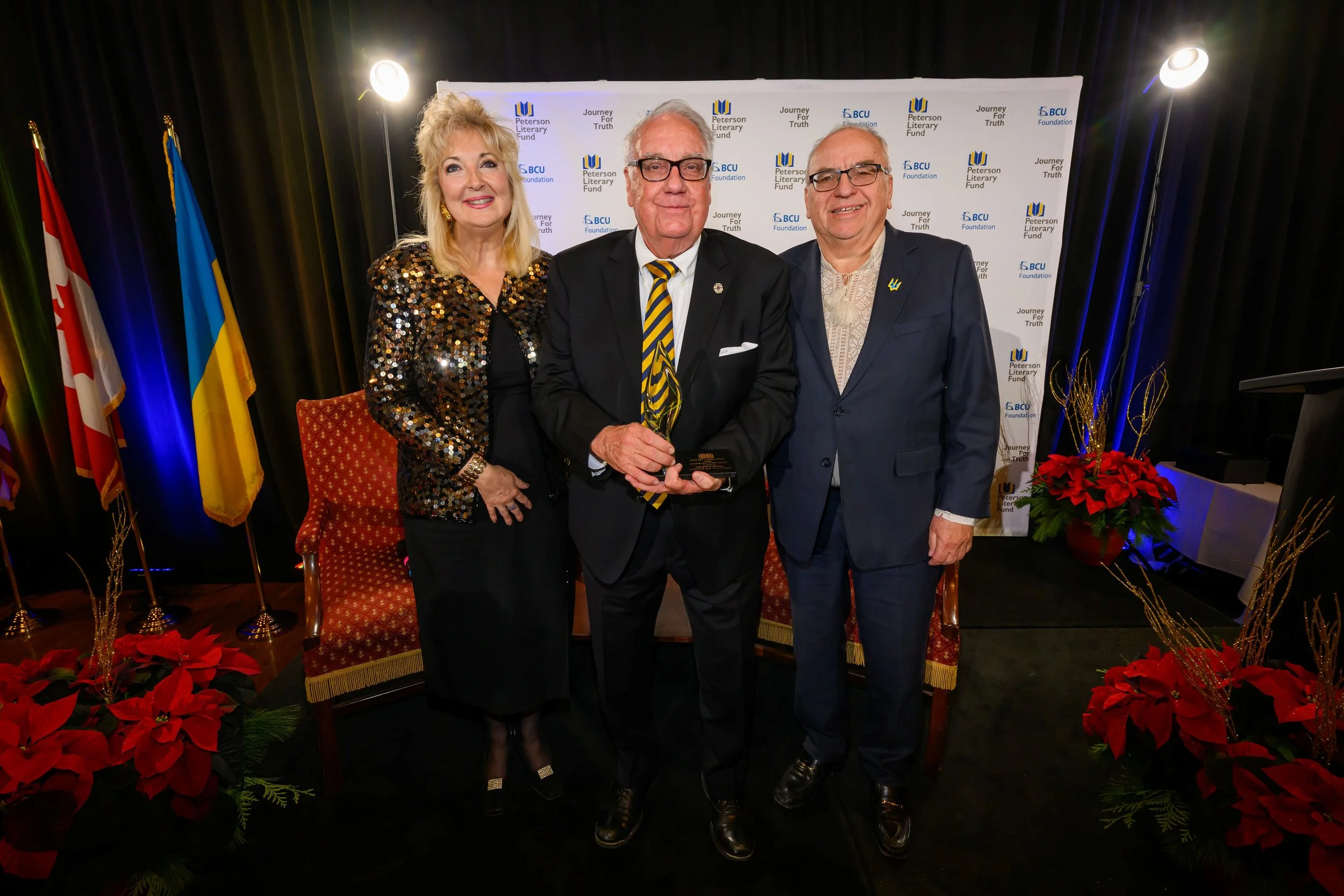 Three people posing at an event in front of a backdrop with logos of Peterson Literary Fund, BCU Foundation, and Journey For Truth. The woman on the left is wearing a sparkly black and gold dress, and the two men are dressed in suits with striped ties. The setting is decorated with flags and red poinsettias.
