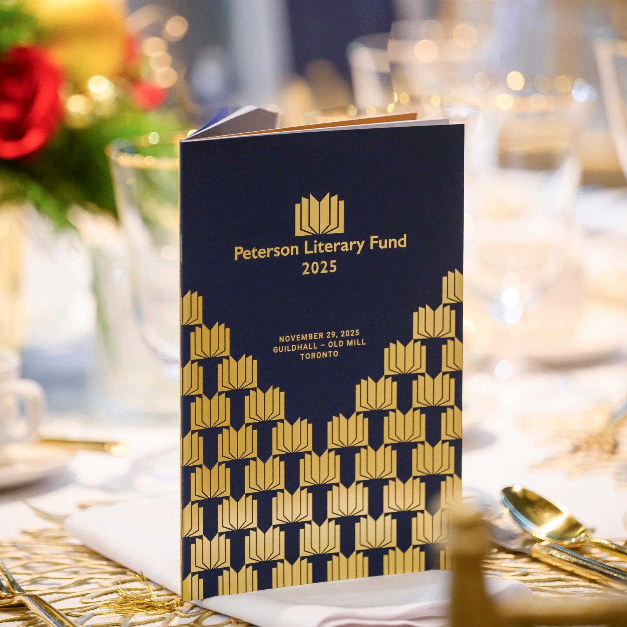 A booklet titled 'Peterson Literary Fund 2025' standing upright on a table with a gold decorative pattern, surrounded by gold confetti and a blurred background with lights and floral arrangements.