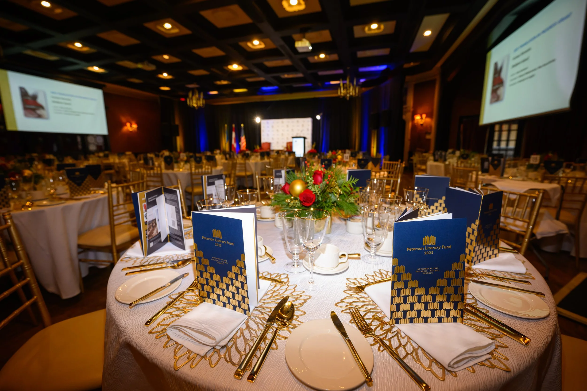 A banquet hall decorated for an event with round tables covered in white tablecloths, adorned with gold utensils, glassware, and black and yellow programs labeled 'Peterson Literary Fund 2025'. The centerpieces feature red roses, gold and red ornaments, and greenery. Large screens are on either side of the stage at the front of the room, which is set with flags and a podium, with dim lighting and chandeliers overhead.