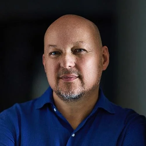 A bald man with a goatee wearing a blue collared shirt, looking at the camera with a neutral expression in a dark indoor setting.