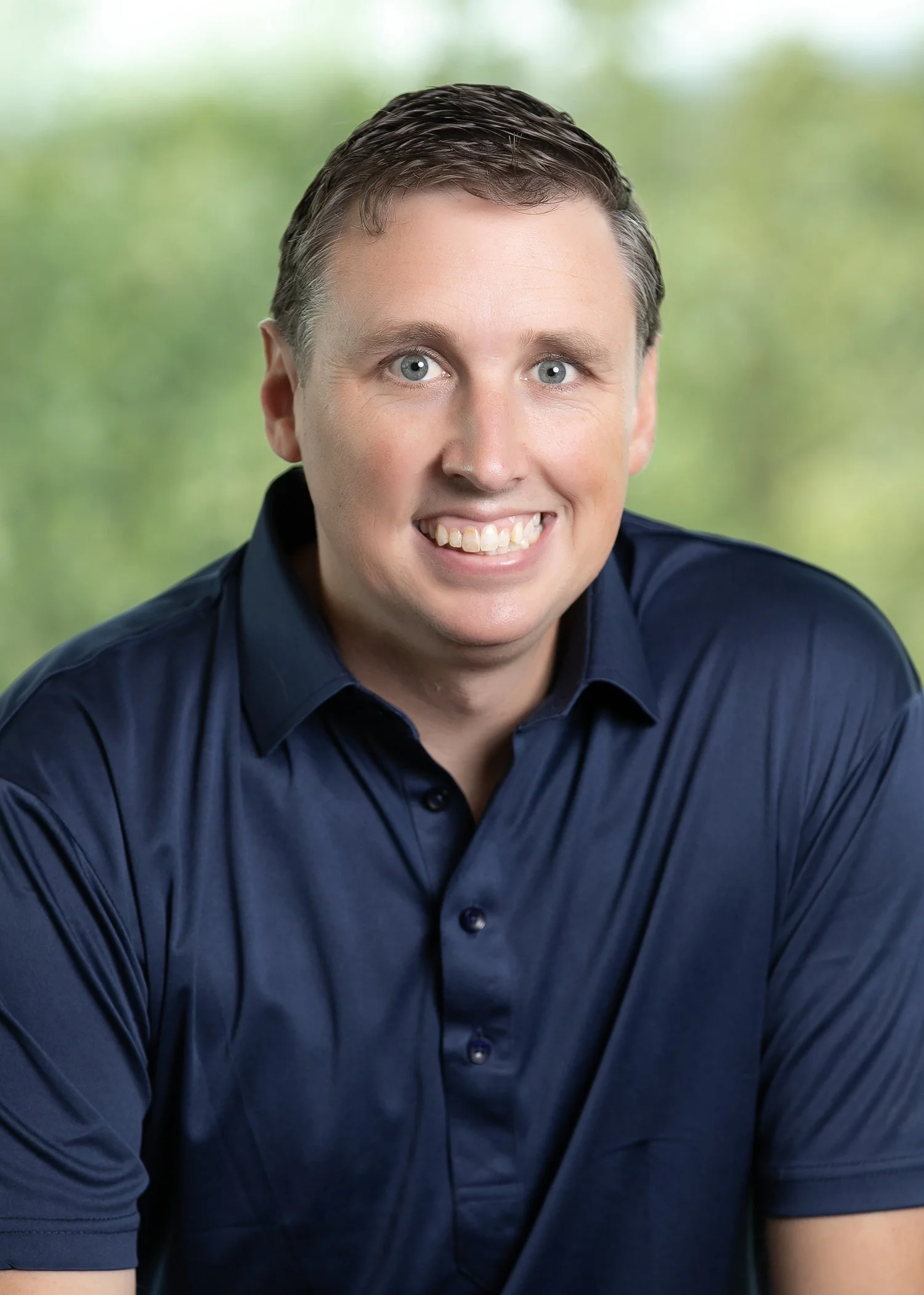 A man with short hair wearing a dark blue polo shirt, smiling against a blurred green background.