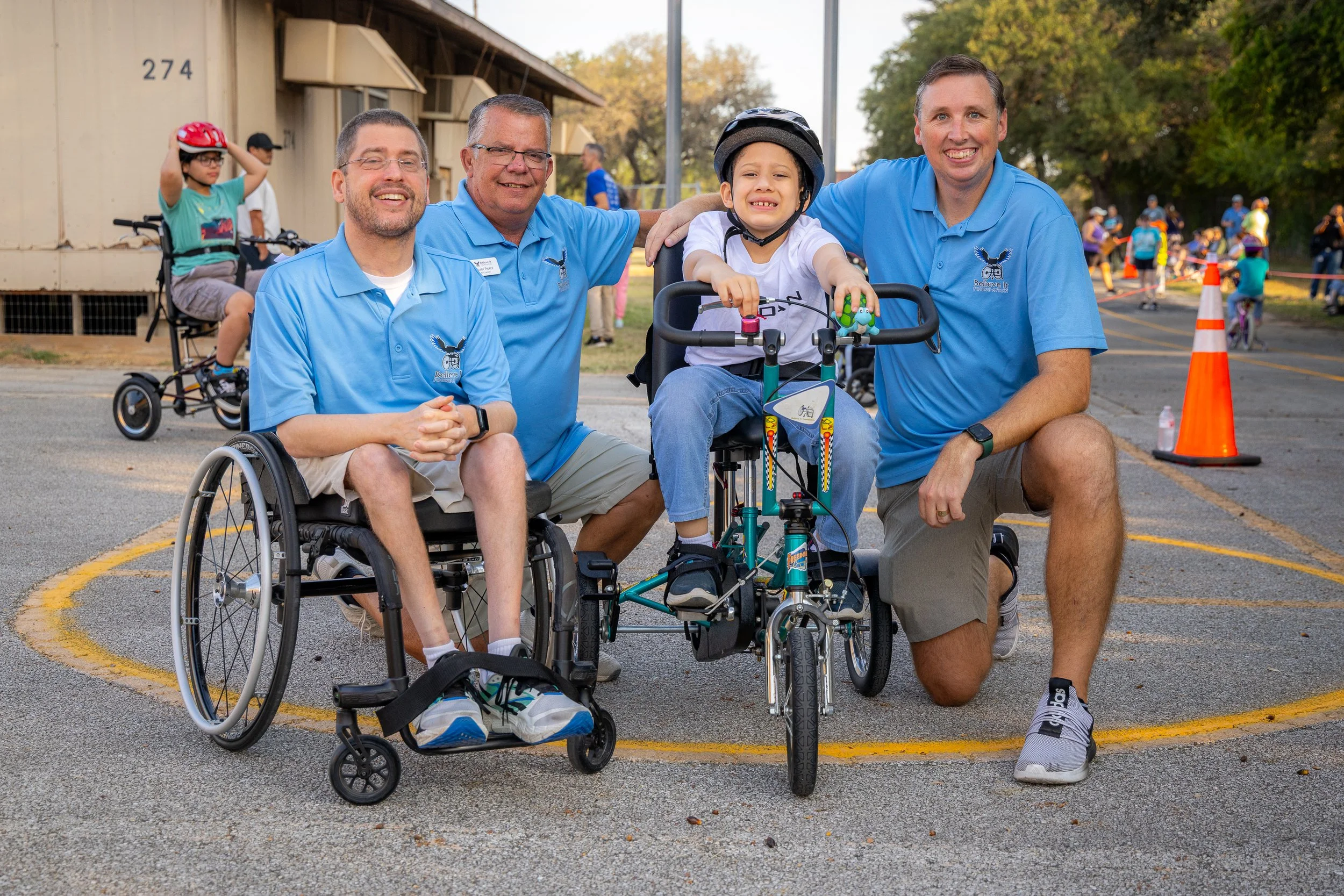 A group of four individuals, including Founder and Staff, smiling outdoors on a paved area with a child with adaptive bike.