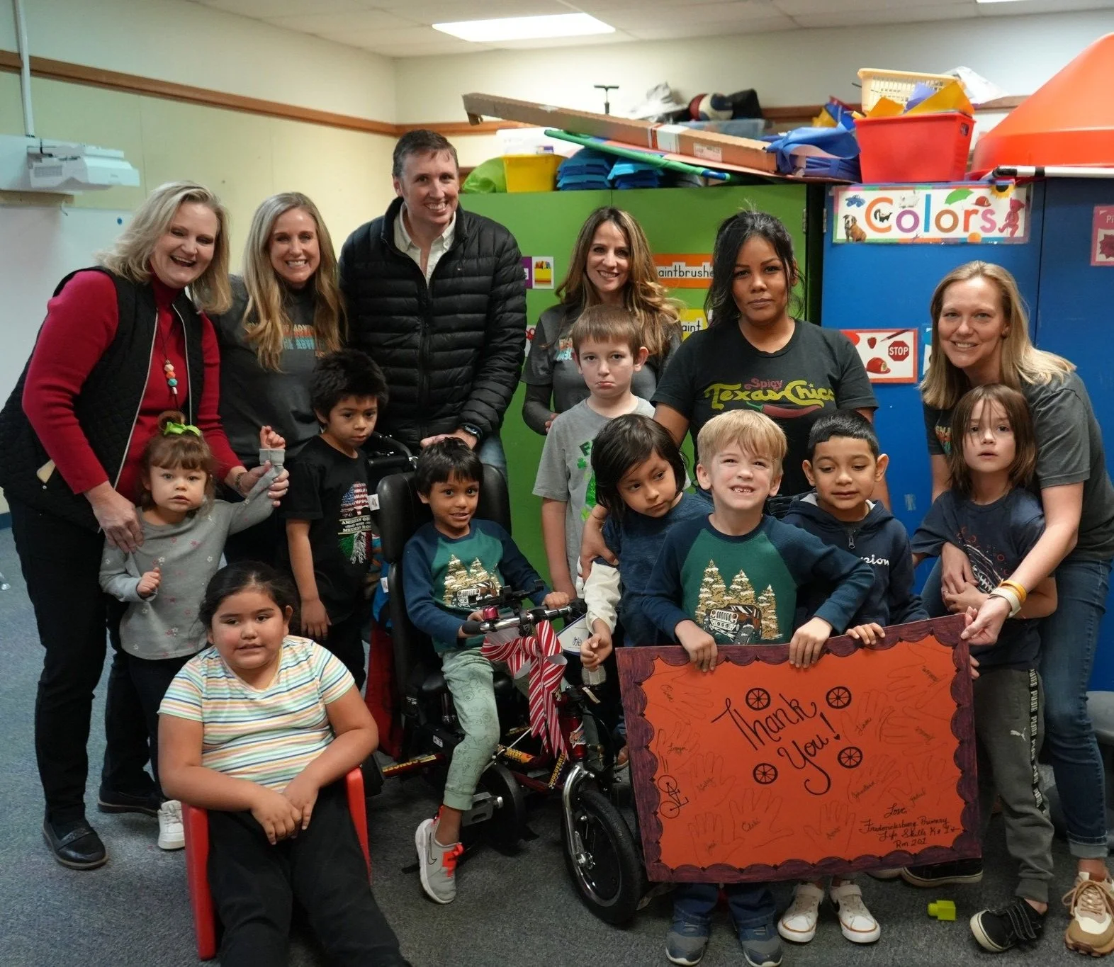 A group of cheerful adult volunteers and children pose together indoors, proudly holding up a hand-painted brown sign that reads "Thank You."