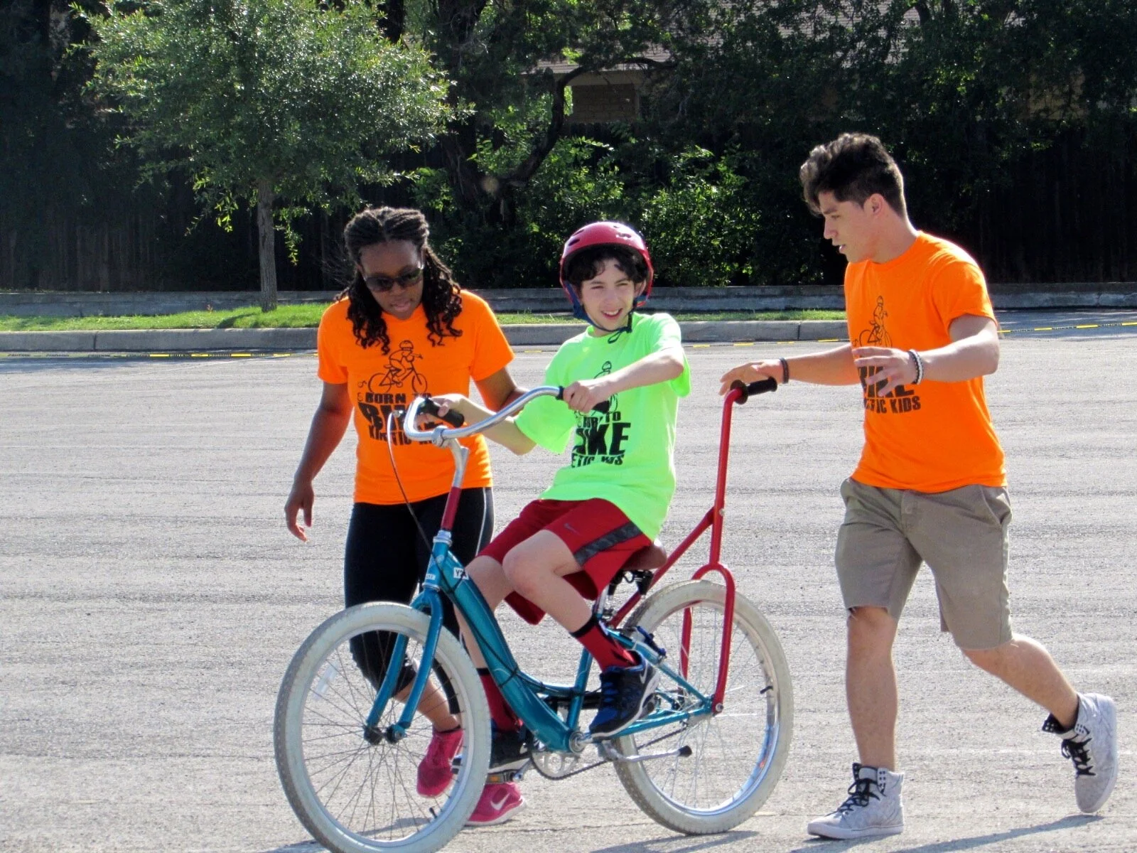 Two volunteer coaches walk alongside a young boy in a helmet, supporting and guiding his blue bicycle on a paved outdoor path.
