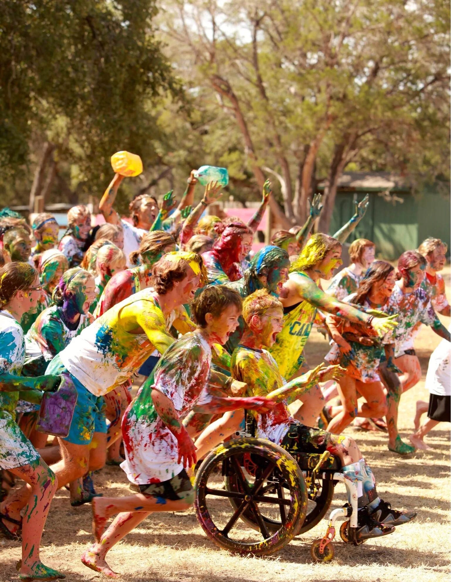 Kids covered in bright paint race outdoors, including a boy in a wheelchair.
