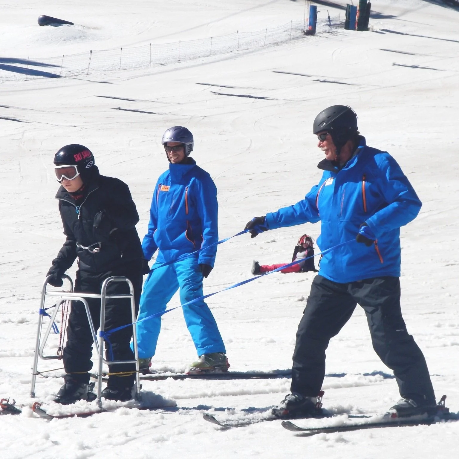 A participant uses a stand-up adaptive skiing frame on a snowy mountain slope, guided closely by an instructor on each side.
