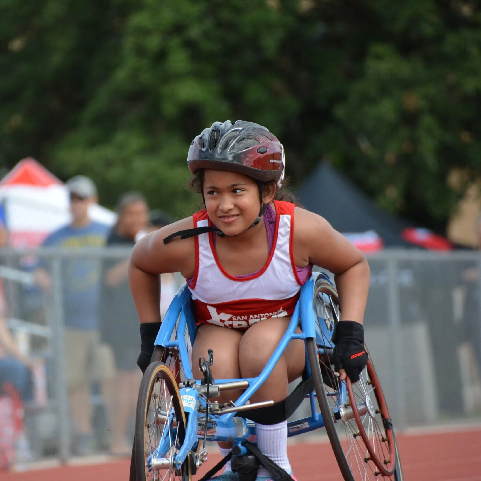 A young female athlete in a helmet and racing uniform pushes a specialized blue racing wheelchair down an outdoor track.