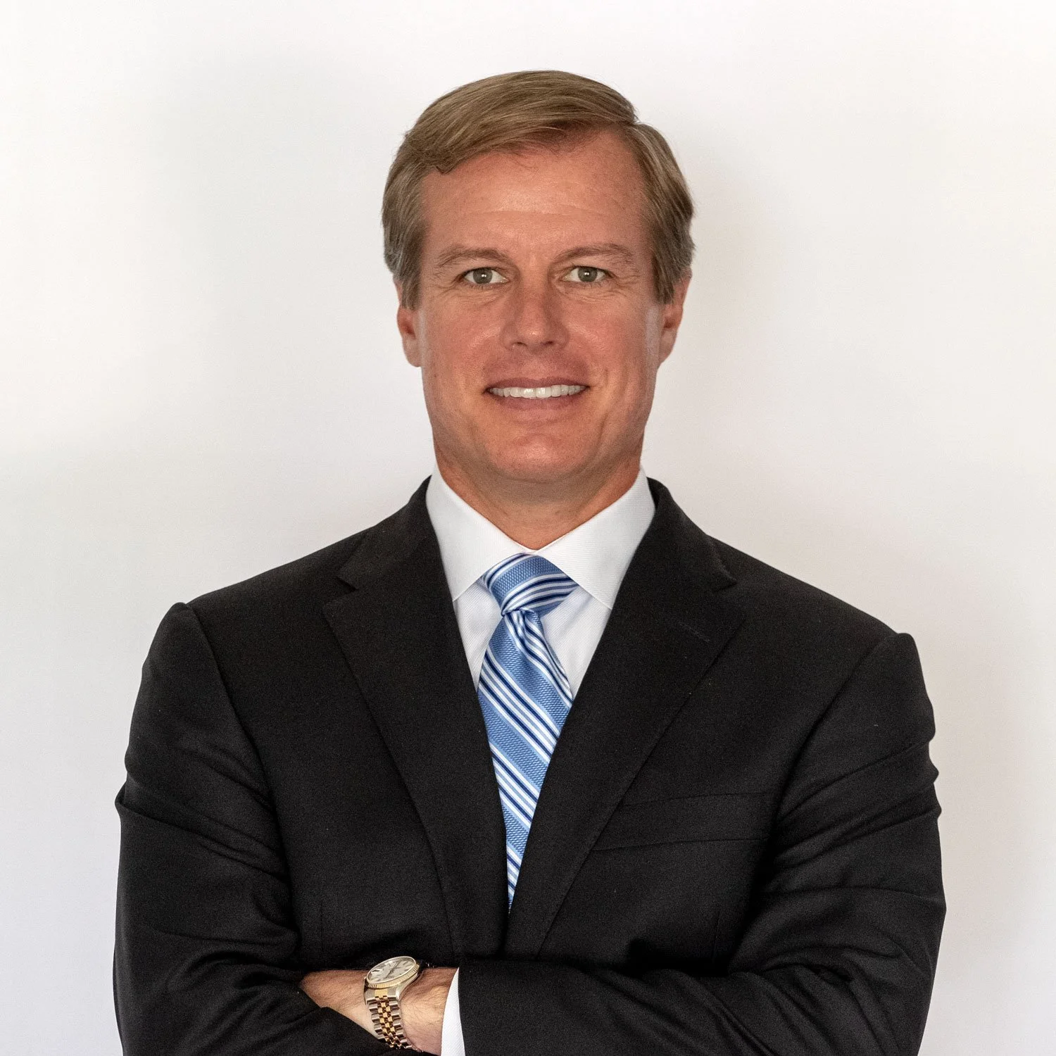 A smiling man with short blonde hair wearing a suit, white shirt, and patterned tie against a light grey background.