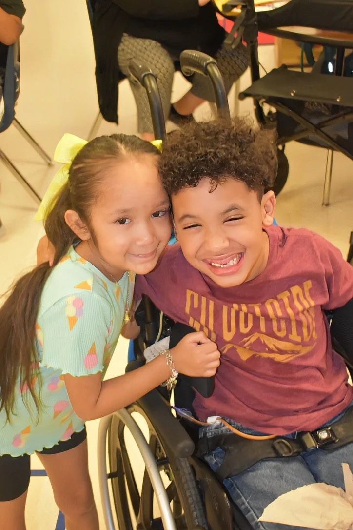 A young girl with a yellow hair bow is smiling while leaning against a smiling boy who is seated in a wheelchair.
