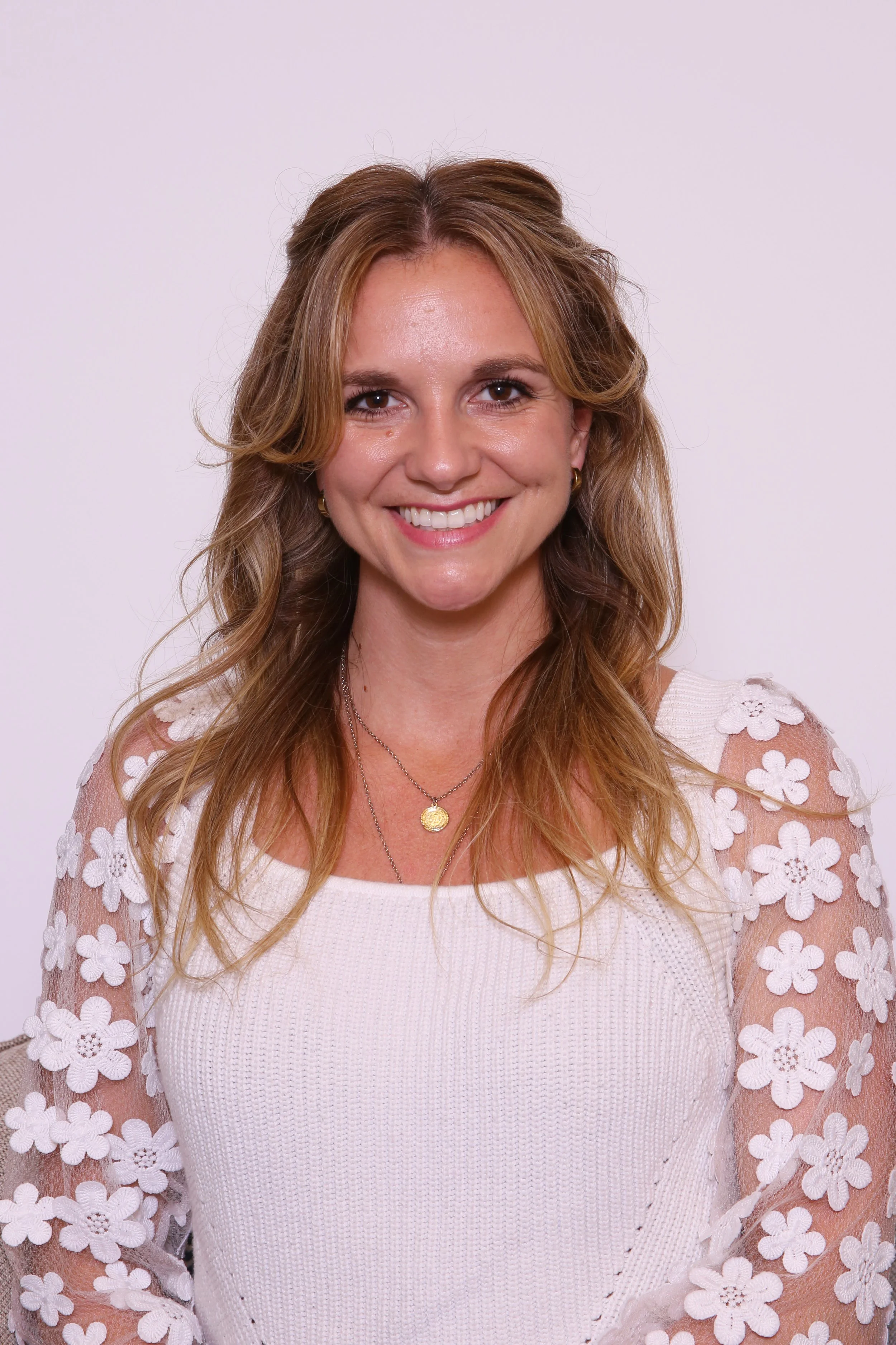 A smiling woman with long wavy blonde hair wearing a white lace top against a lavender background.