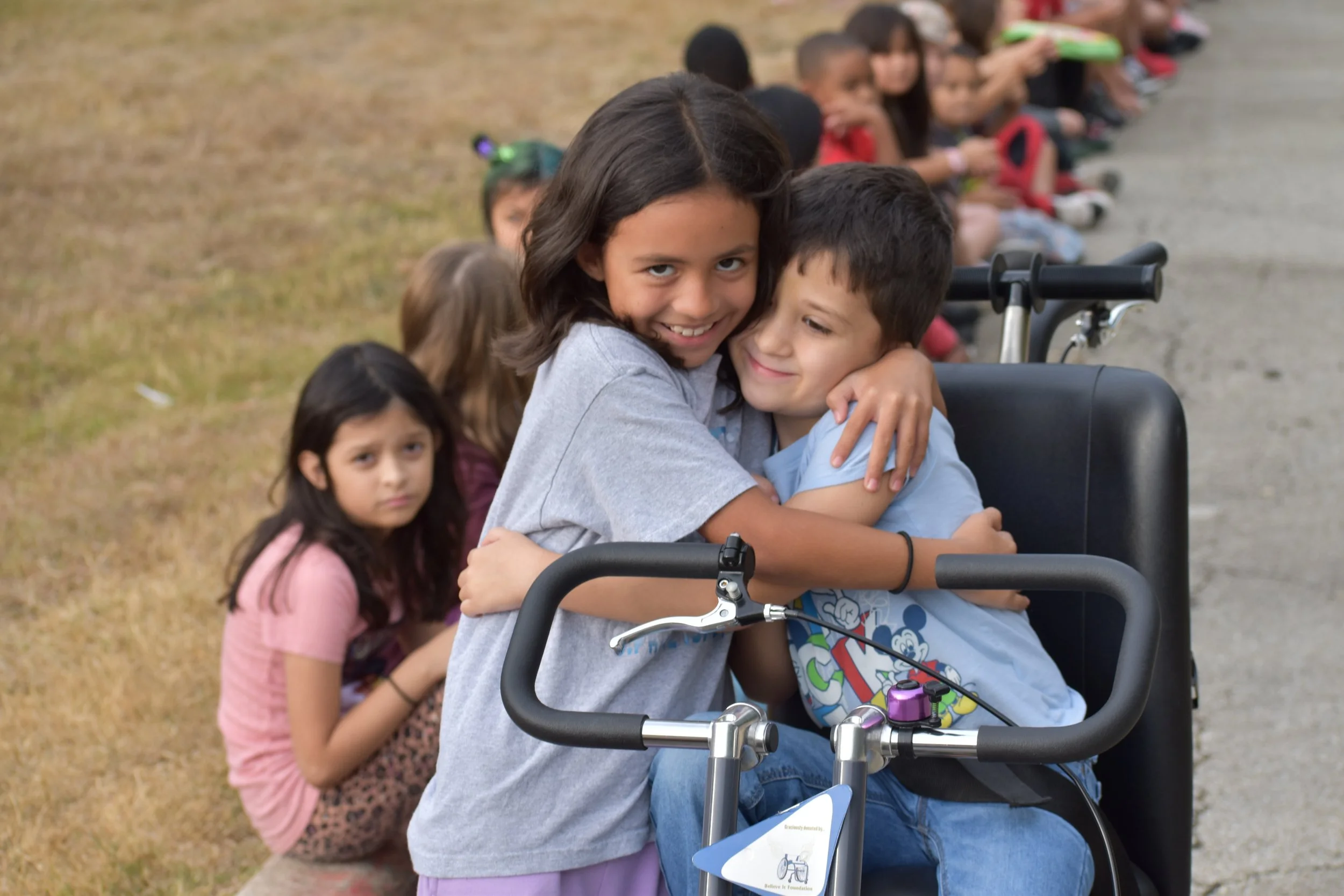Two young friends share a warm hug next to an adaptive tricycle.