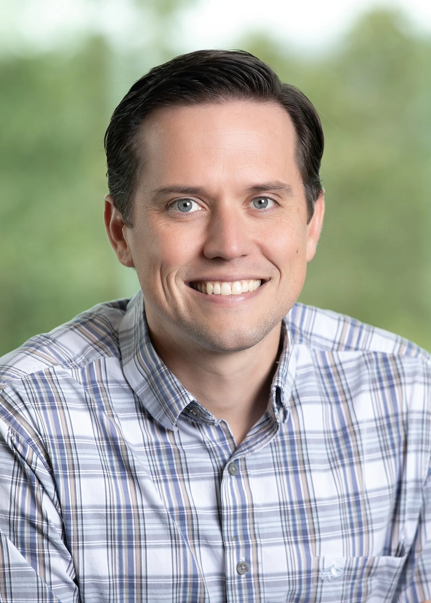 A smiling man with short styled hair wearing a blue and grey plaid button-up shirt against a light background.
