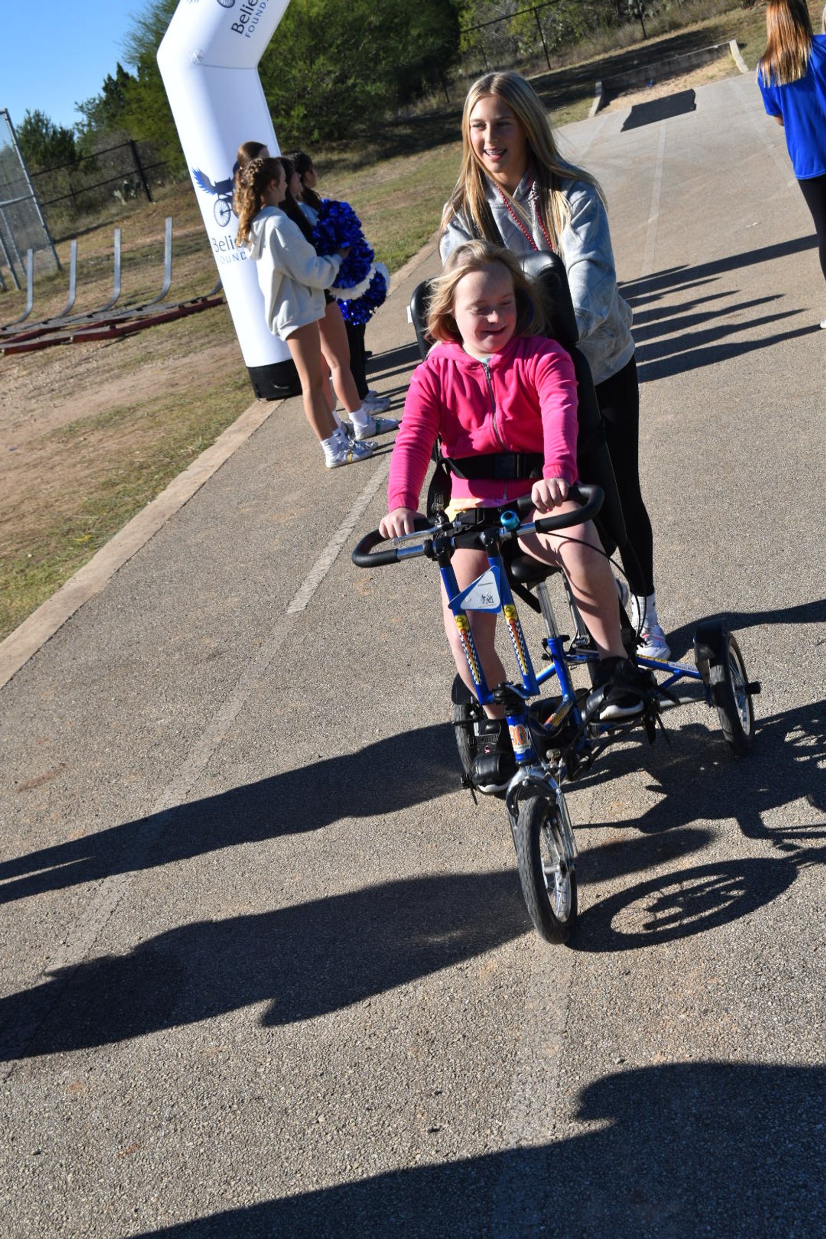 A smiling girl rides an adaptive tricycle past a race finish line.