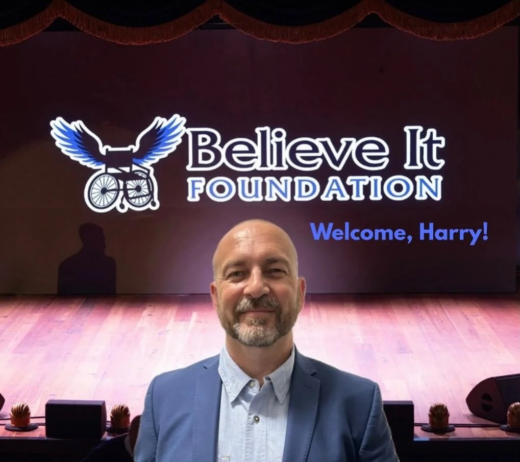 A smiling man in a suit jacket standing on a stage in front of a digital Believe It Foundation logo.
