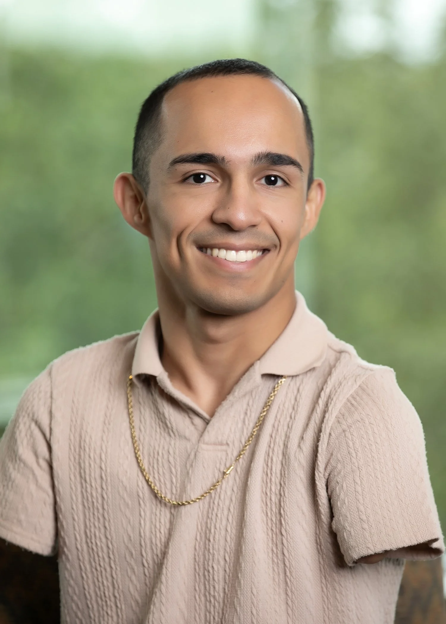 A smiling man with a light beard wearing a textured tan polo and gold chain in an outdoor setting.