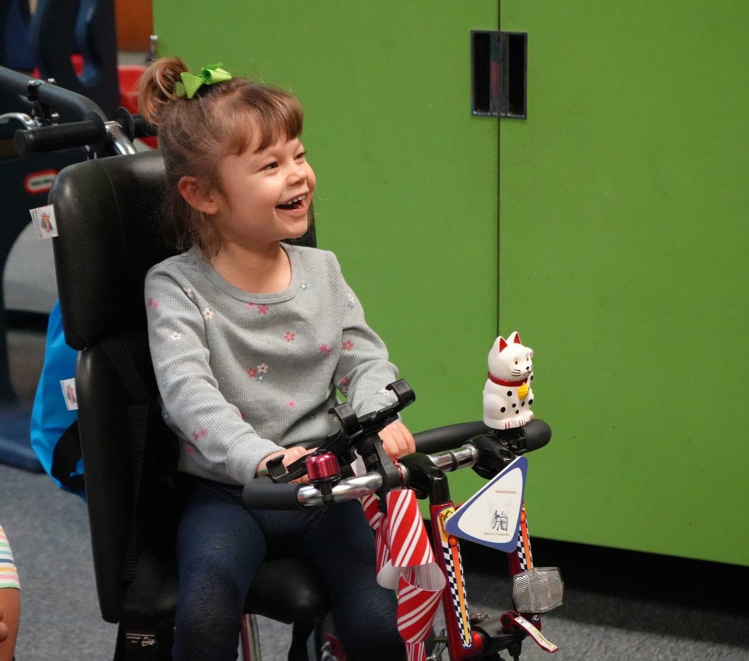A young girl smiles joyfully while seated in a custom, supportive mobility chair equipped with specialized handlebars and toys.