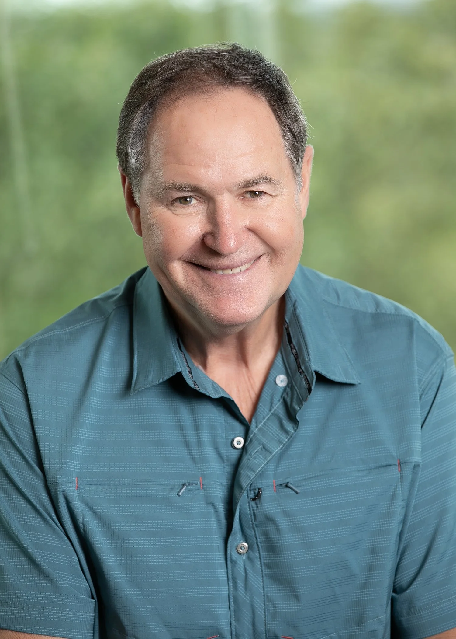A smiling man with short dark hair wearing a casual teal button-up shirt in an outdoor setting.