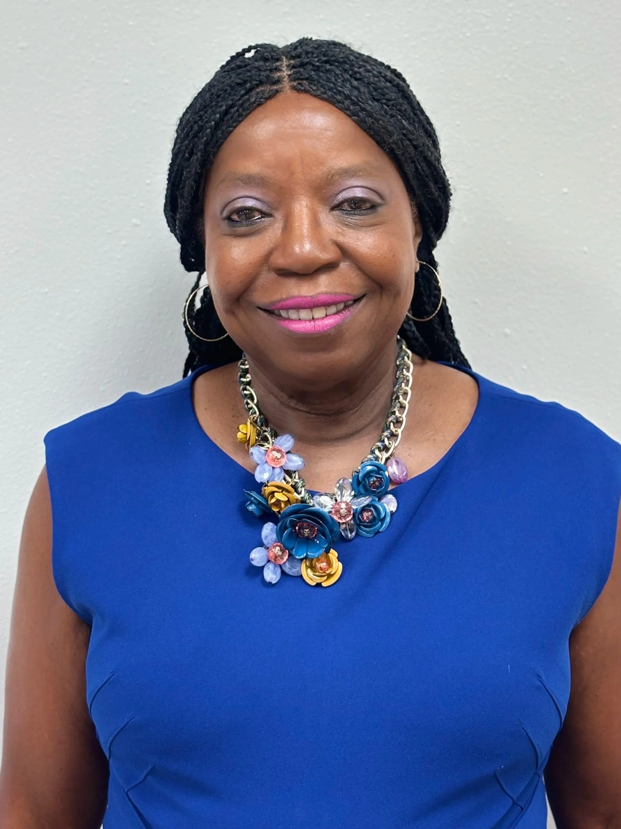 A smiling woman with dark braided hair wearing a royal blue top and a statement necklace against a neutral background.