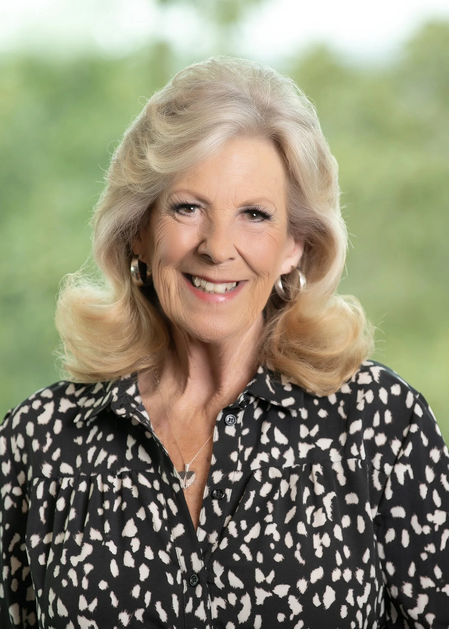 A smiling woman with blonde layered hair wearing a black-and-white patterned top in an outdoor setting.