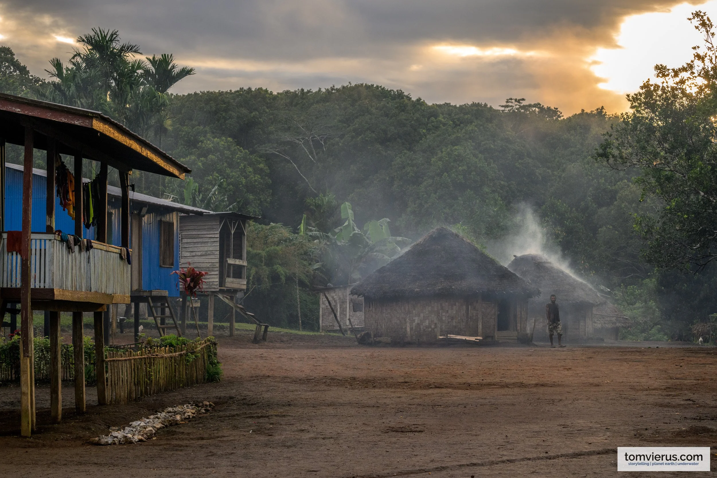 Early morning in Pakia village, New Britain, with smoke rising between thatched houses and sunlight breaking through clouds above forested hills.