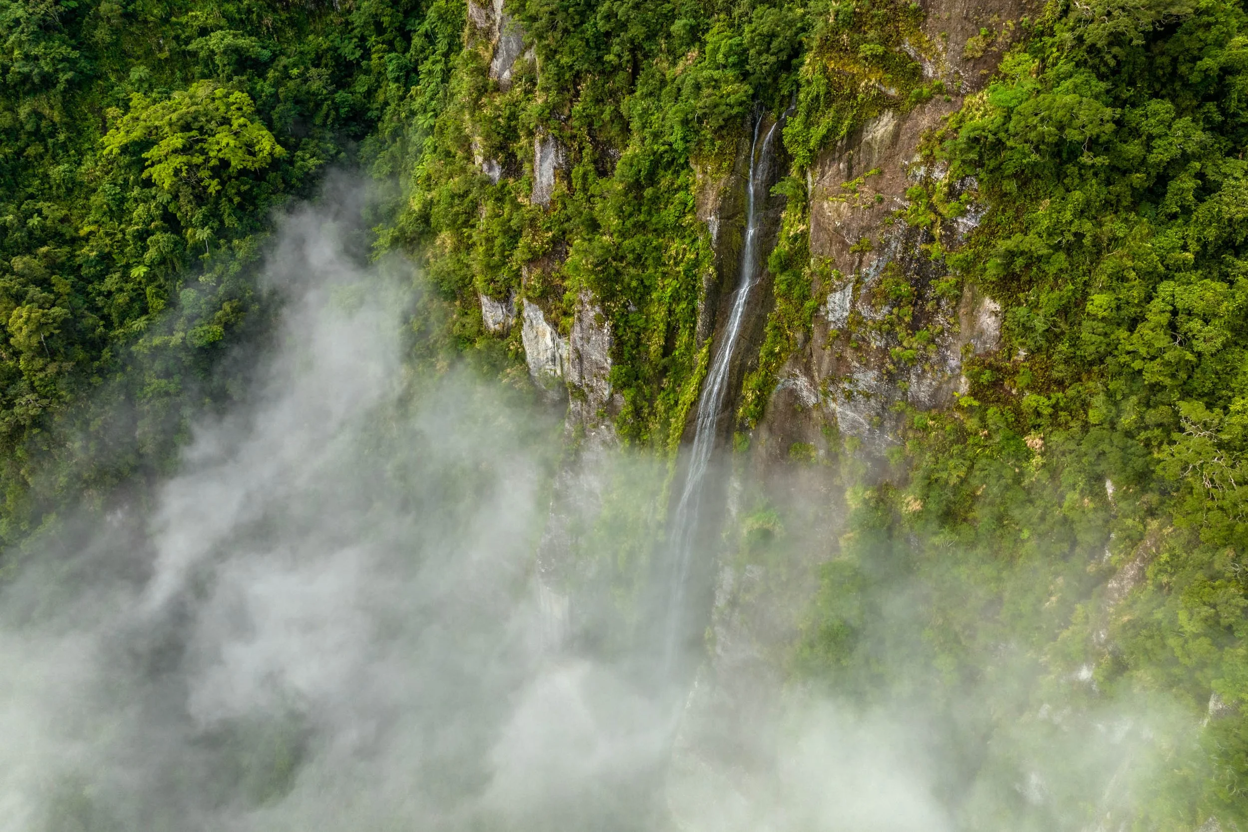 Aerial view of a tall, thin waterfall cascading down a lush green mountainside with fog near the base.
