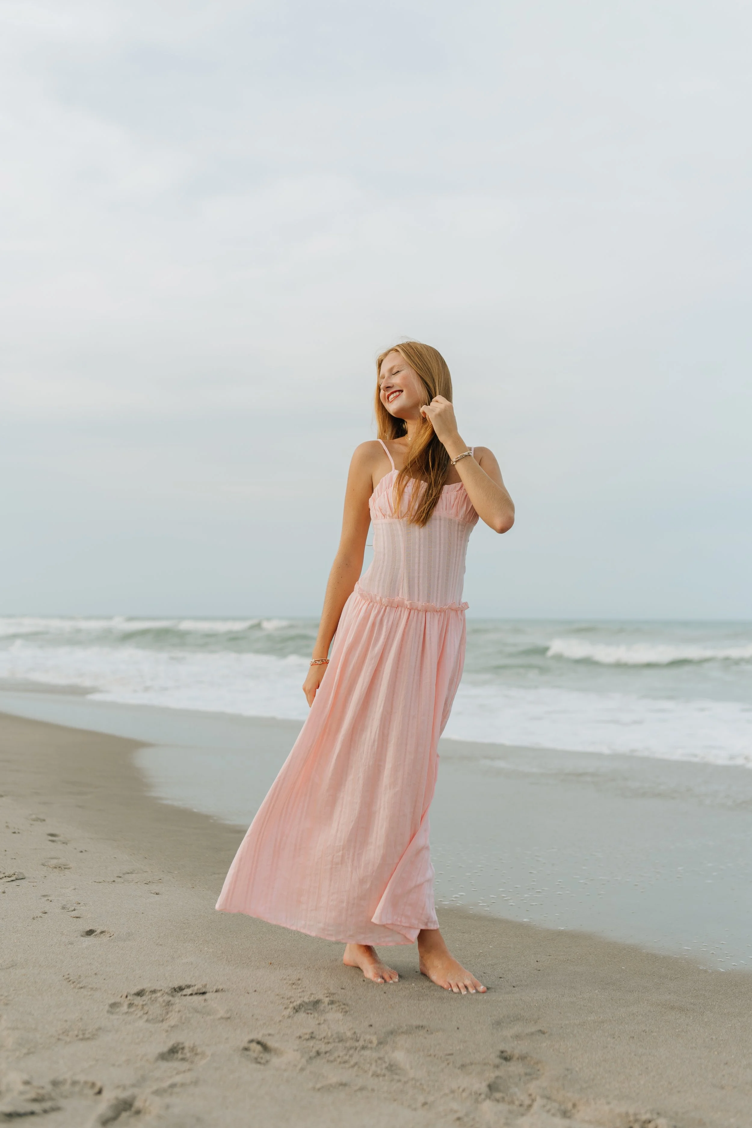 A high school senior girl on the beach at sunset during her senior session in a pink flowy dress.