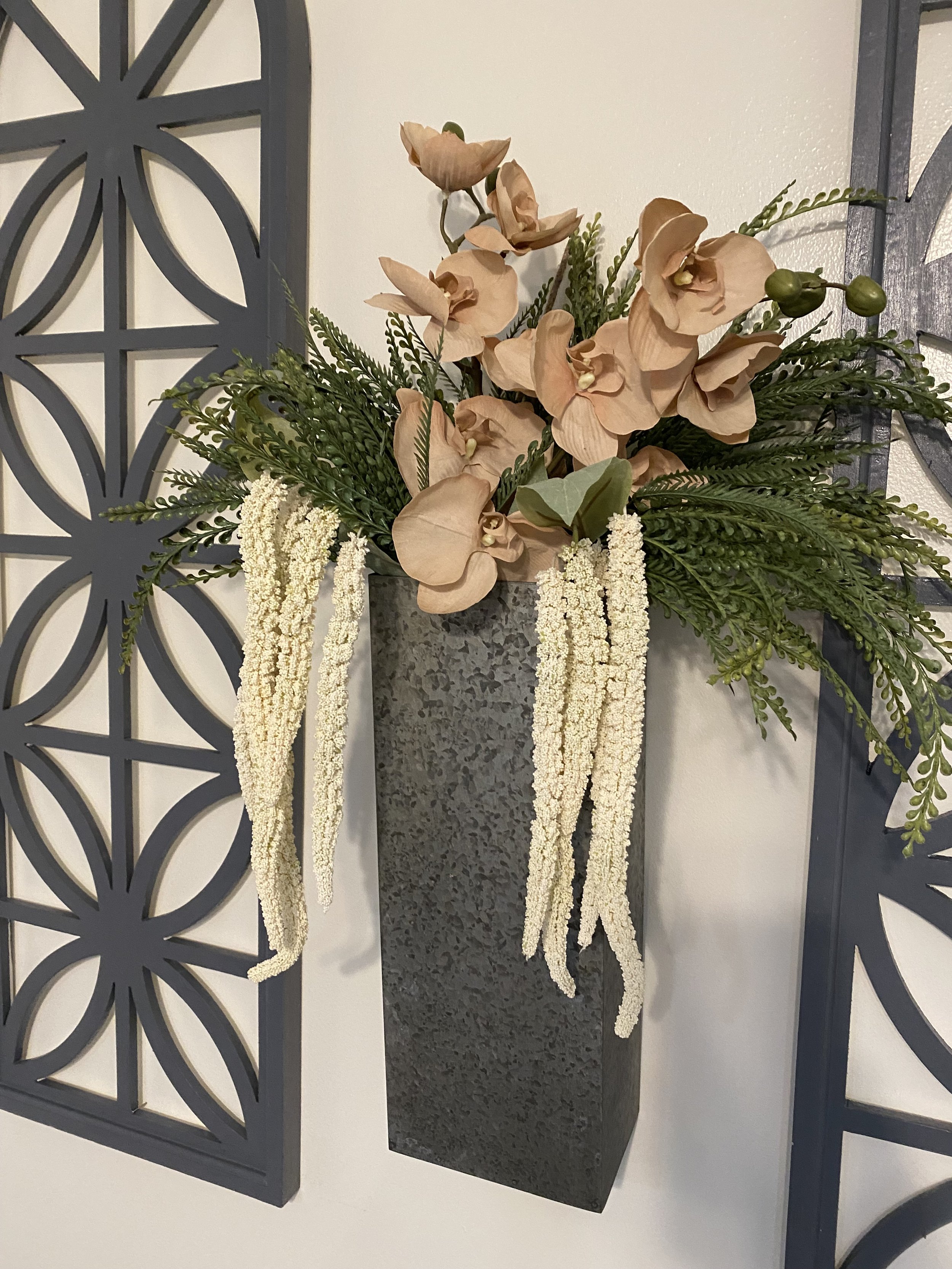 Arrangement of artificial peach-colored orchids, green foliage, and white hanging accents in a tall, textured gray vase on a wall with black metal decorative panels.