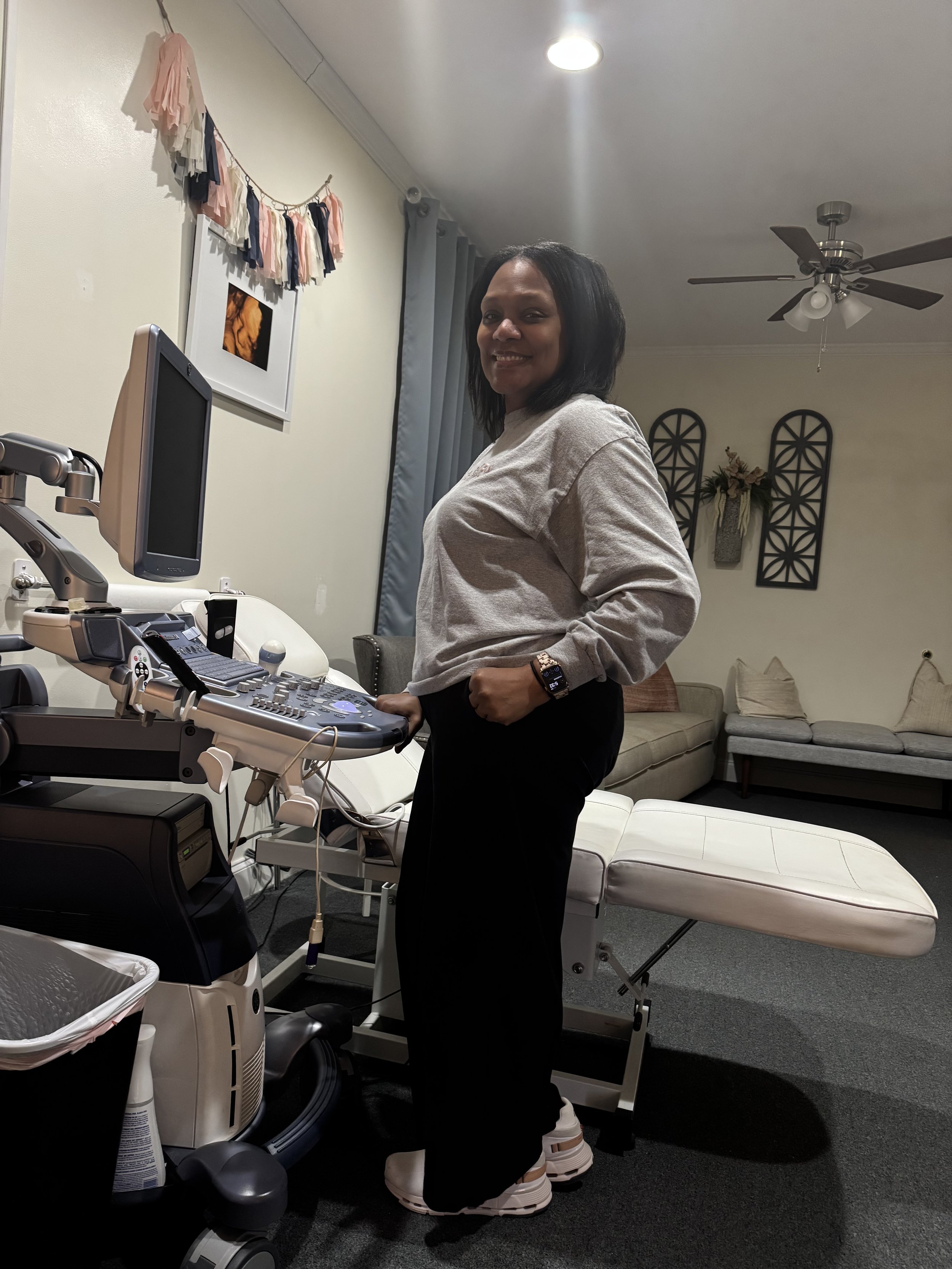 A woman standing beside an ultrasound machine in a room with a cream-colored couch, wall decorations, and a ceiling fan.
