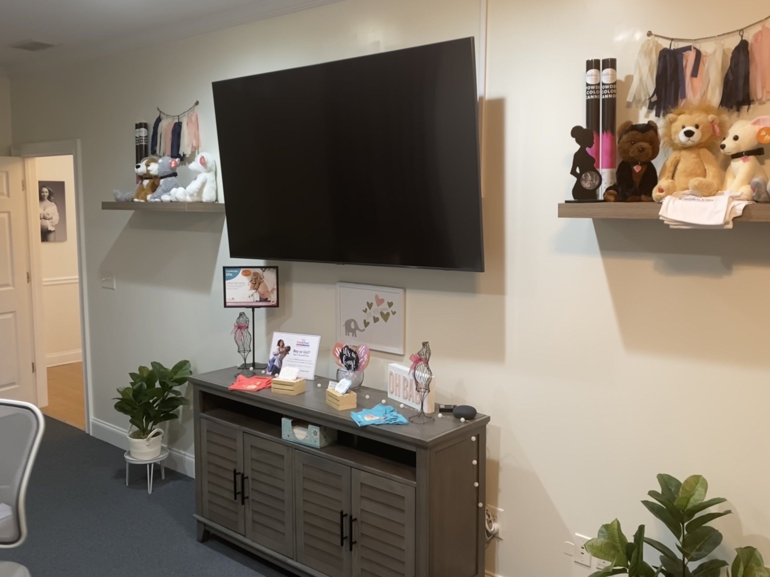 Living room with a wall-mounted flat-screen TV above a gray wooden cabinet, decorated with plush toys on floating shelves, people figurines, and framed pictures, with a potted plant and a white chair nearby.