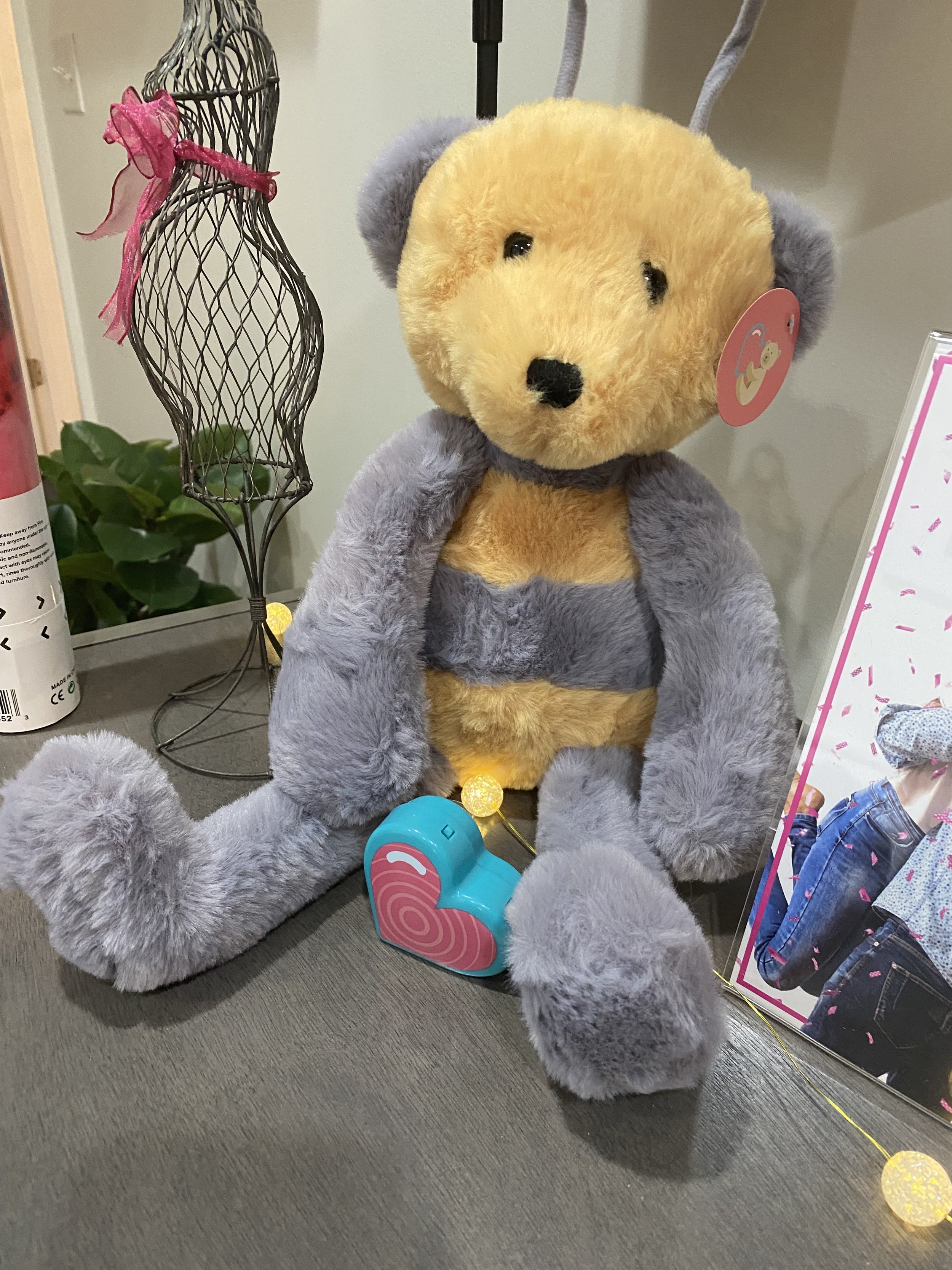 A plush heartbeat teddy bear with a yellow face, gray ears, and gray arms, sitting on a dark surface near a decorative wire mannequin and a card with a photo of a person in blue jeans.