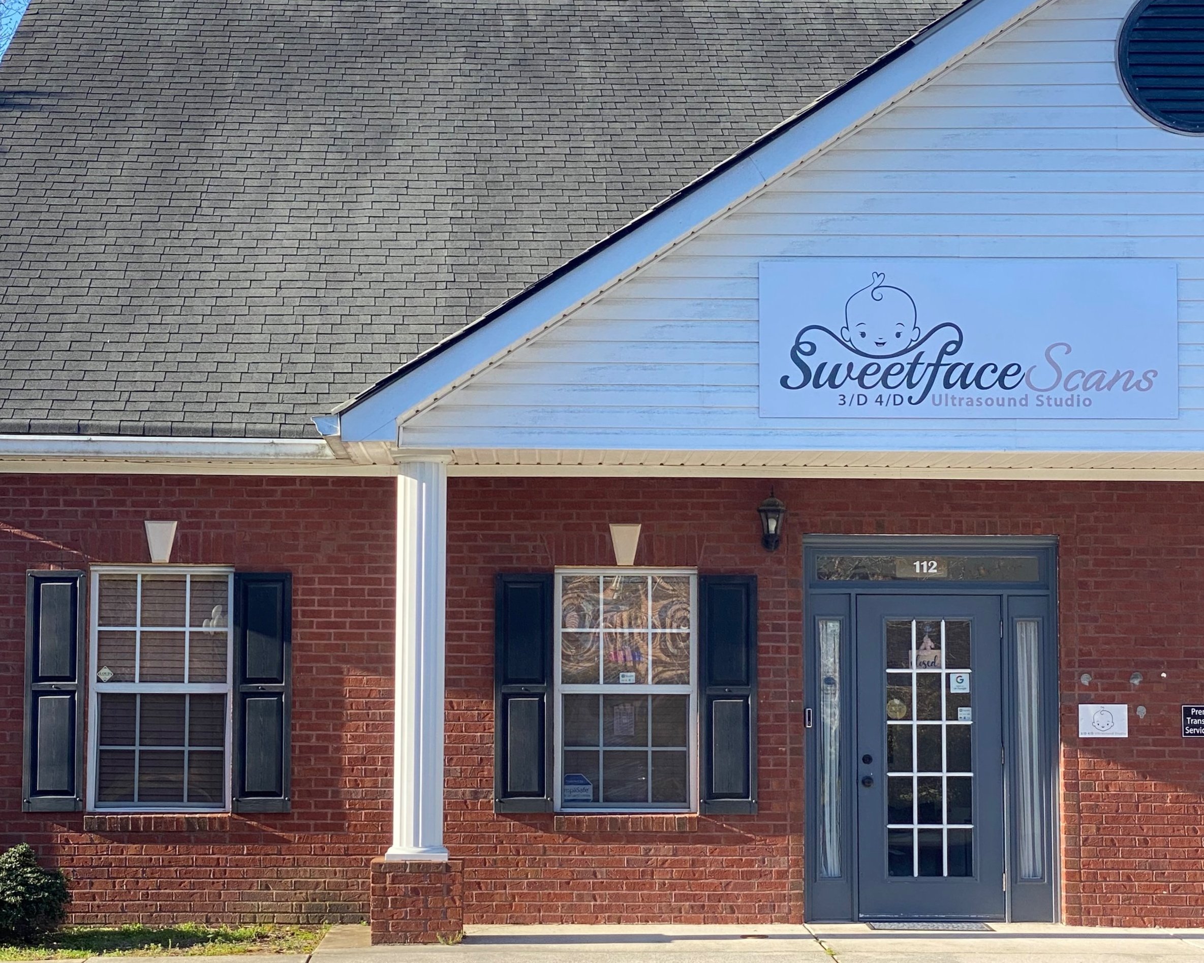 Brick building housing Sweetface Scans, a 3D and 4D ultrasound studio, with a sign featuring a baby face illustration and the studio's name above the entrance, which has a glass door.