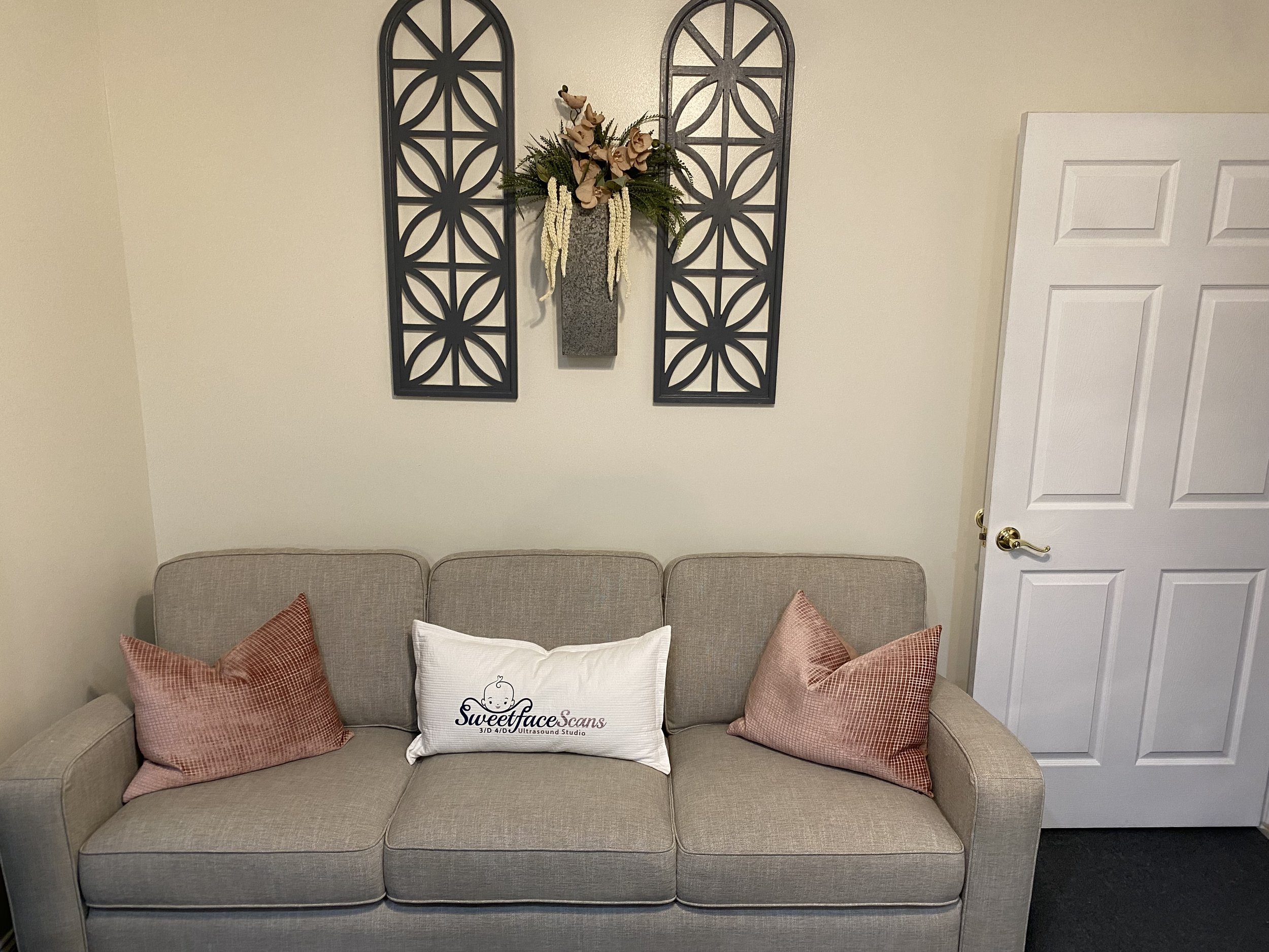 A beige sofa with two pink textured pillows and a white pillow that reads 'Sweetface Scans Ultrasound Studio.' Above the sofa are two decorative black metal wall panels and a wall-mounted vase with flowers and greenery.
