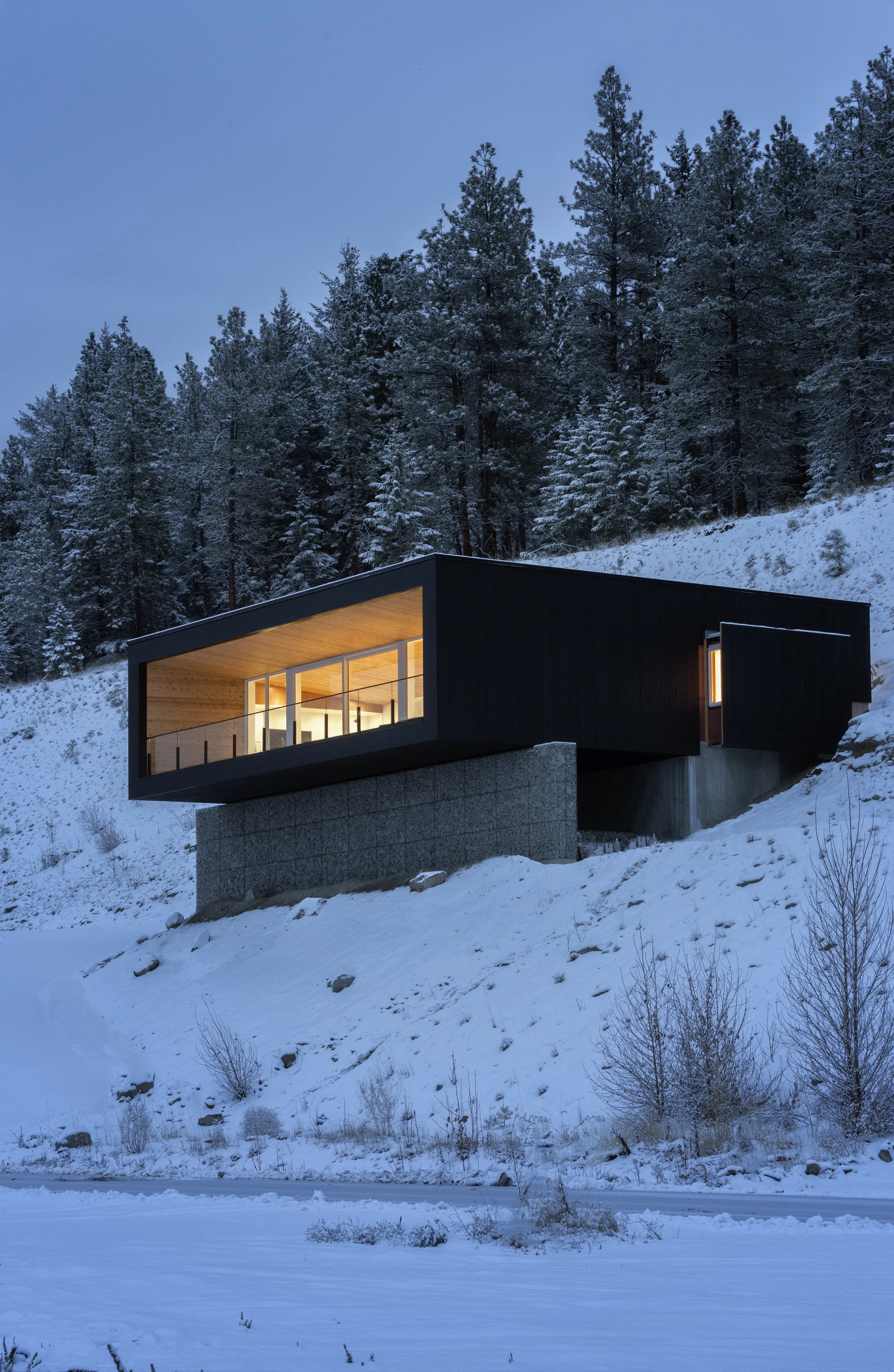 Modern house on a snowy hillside surrounded by trees, with built-in lights illuminating the interior, visible through large windows.