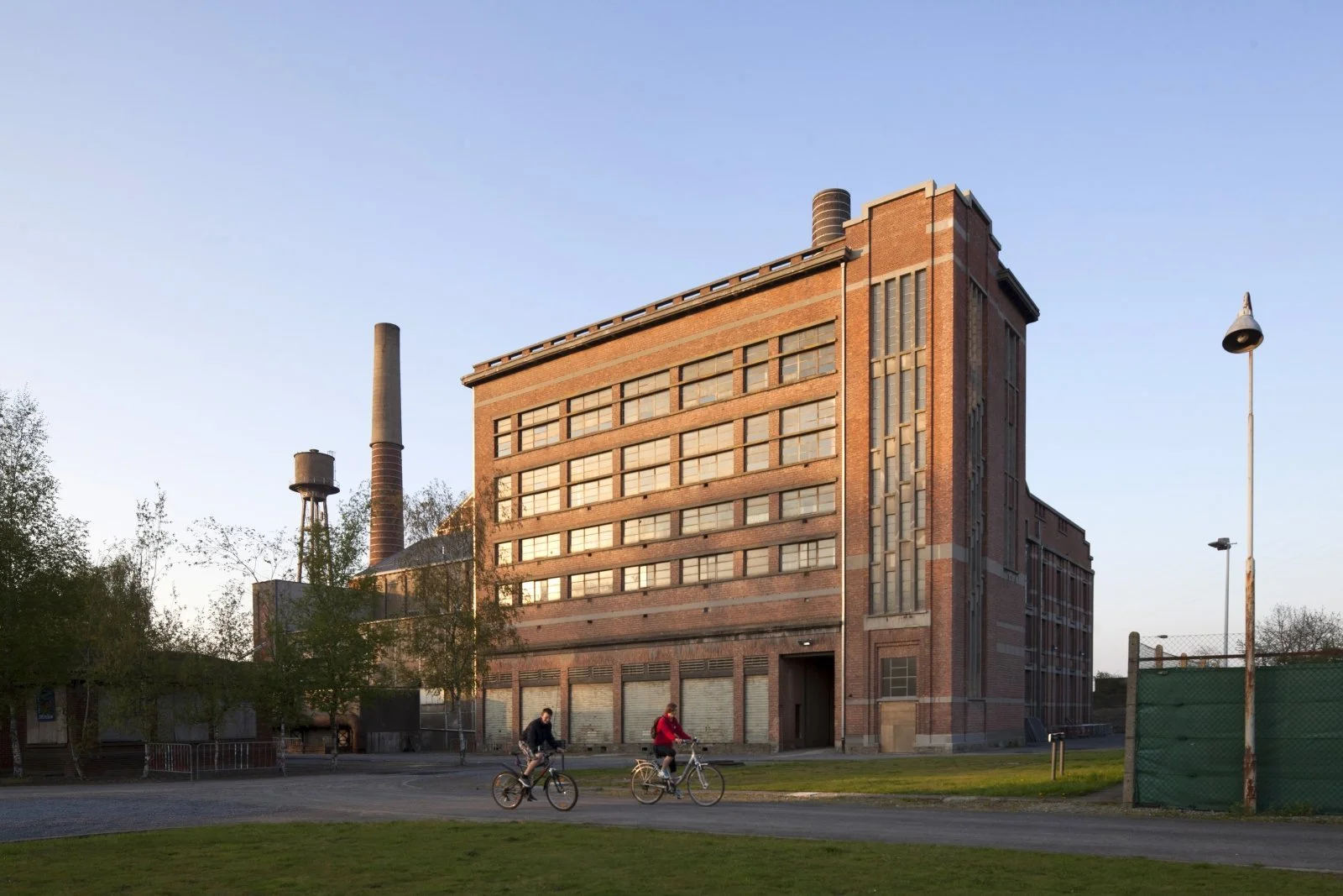 Edificio industrial de ladrillos, rehabilitado con bloques de hormigon de cañamo, con chimeneas altas, dos personas pedaleando en bicicletas en primer plano, área verde y árbol, cielo despejado.