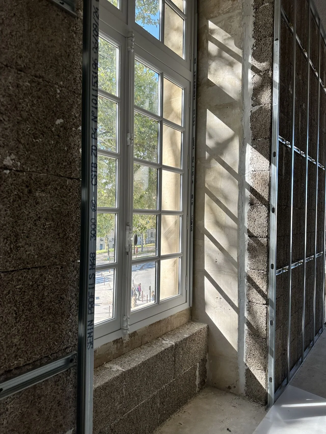 Ventana de doble hoja en construccion, con marco de PVC blanco y cristales transparentes, en una pared de ladrillos de hormigon de cañamo hempcrete, con sombra en la pared y vista de arboles y personas afuera.