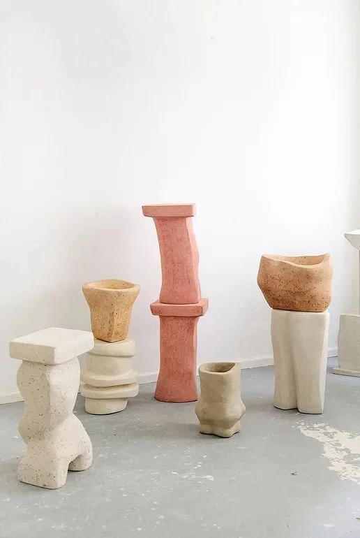 Esculturas modernas de forma abstracta, fabricadas en hormigon de cañamo, en tonos blancos, beige, rosa y marrón en una galería con paredes blancas y suelo de cemento.