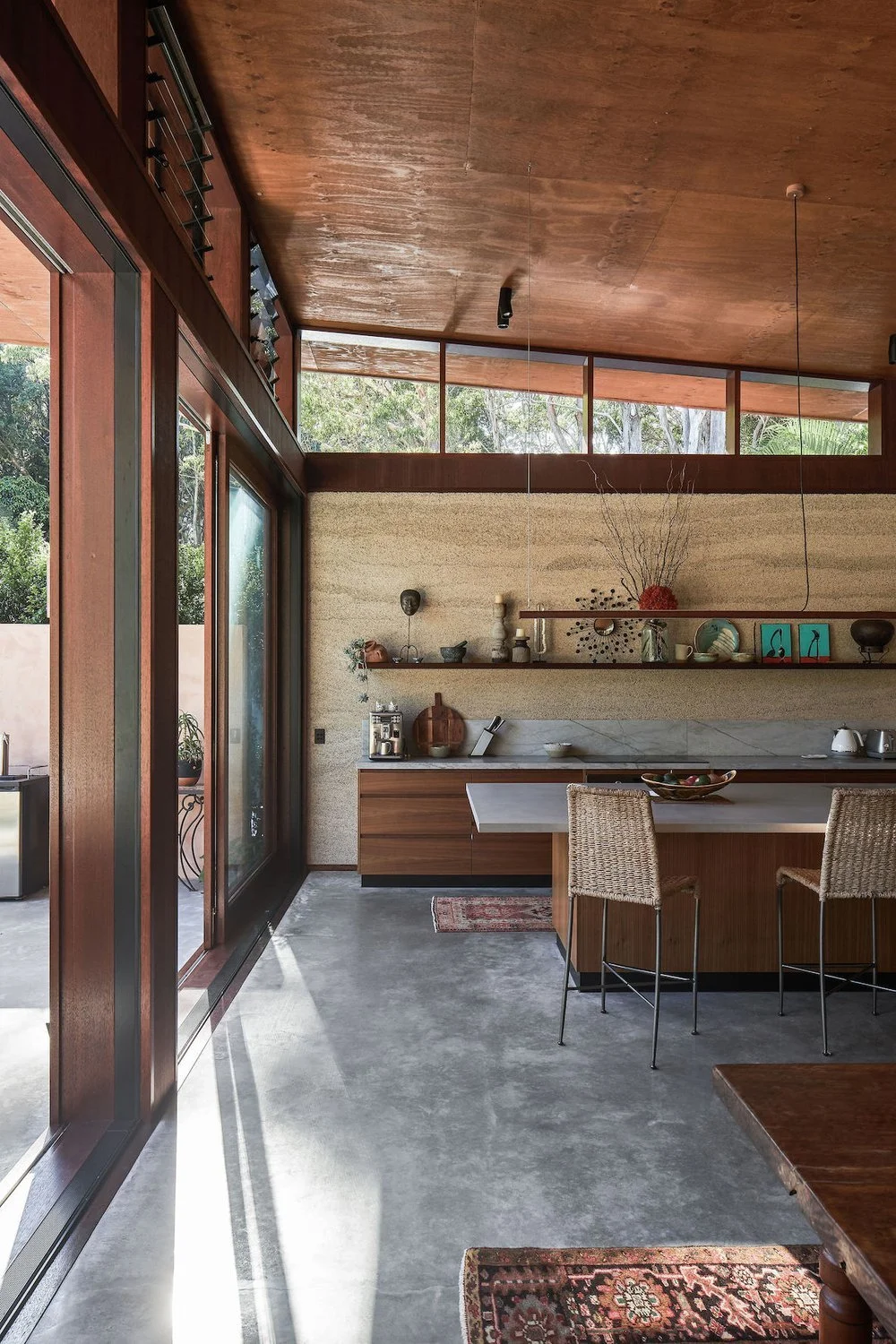 Cocina moderna con paredes de tapial de hormigon de cañamo, mobiliario de madera, pisos de concreto, y ventanas grandes que dejan entrar luz natural, con una mesa y sillas de mimbre.